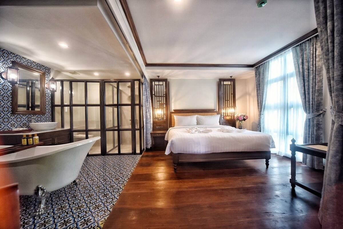 Une chambre spacieuse avec un grand lit, une salle de bain séparée avec une baignoire et une grande fenêtre avec des rideaux.
