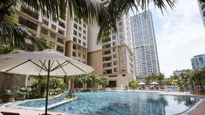 A swimming pool with lounge chairs and umbrellas, surrounded by palm trees and a tall building in the background.