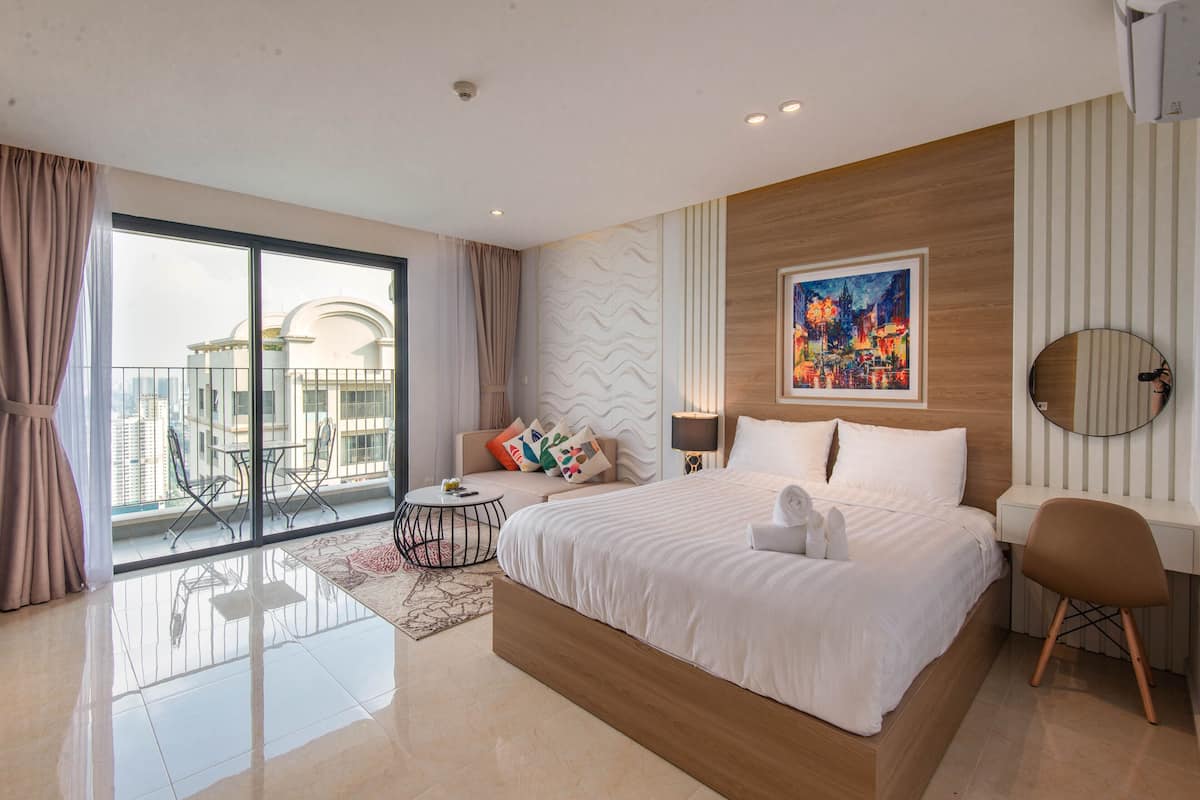 A modern hotel room with a large bed, a desk, a chair, a round mirror, a painting, and a balcony with a city view.