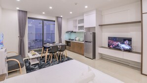 A modern hotel room with a bed, a dining area, a kitchen, and a TV displaying a concert.