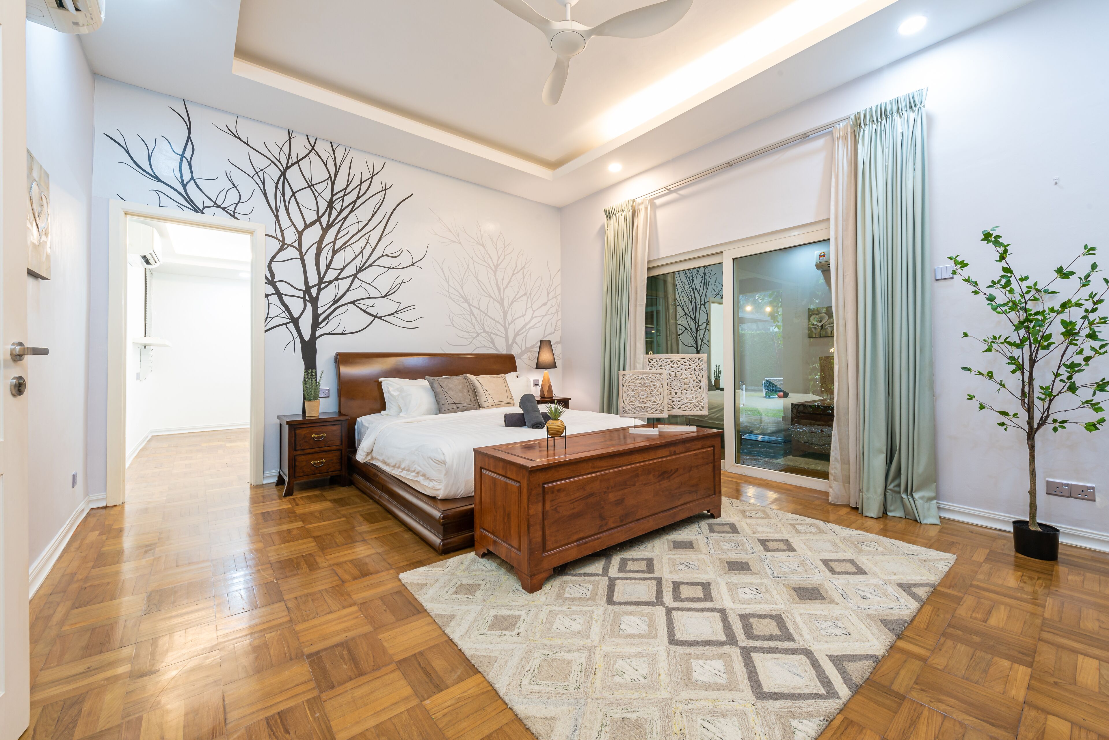 A bedroom with a wooden bed, a nightstand, a dresser, a large window with curtains, and a potted plant.