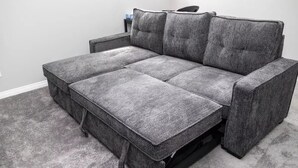Ein graues Ecksofa mit passendem Hocker in einem Raum mit grauem Teppich und einem kleinen Beistelltisch.