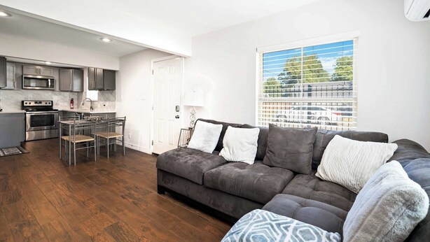 A modern living room with a dark grey sectional sofa, a dining area with a metal table and chairs, a kitchen with stainless steel appliances, and a large window with blinds. - Texas Med Center Stunning & Vibrant Stay (Houston)