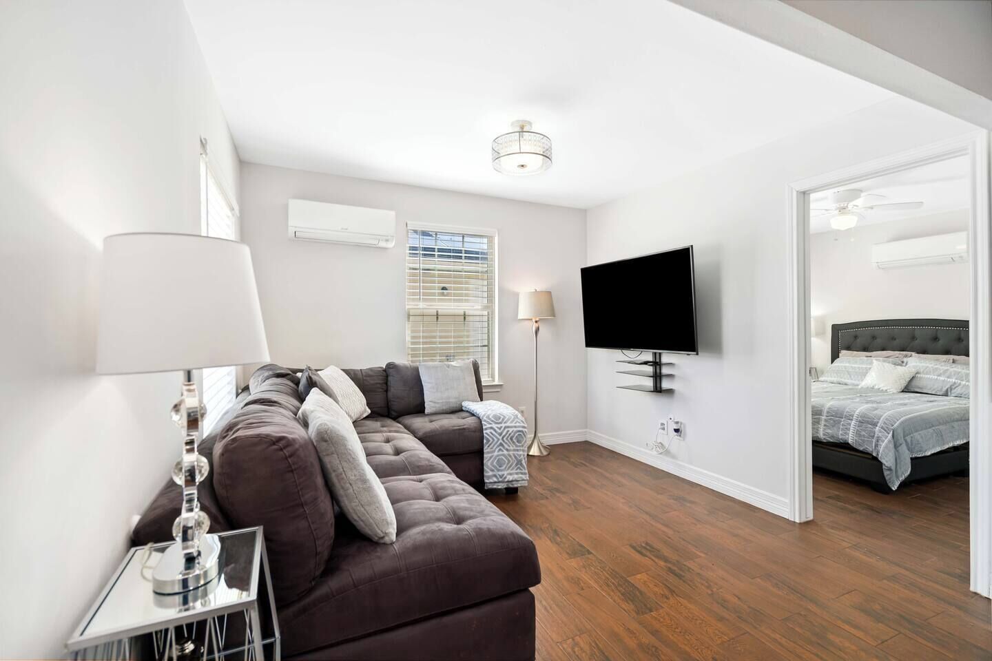 A modern living room with a sectional sofa, a flat-screen TV mounted on the wall, a standing lamp, and a bed with a dark headboard.