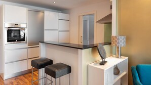 A modern kitchen with a built-in oven, two black bar stools, a decorative lamp, and a blue chair. - Apartment with great sea views on the beach (Marbella)