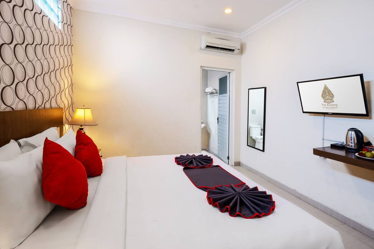 A hotel room with a bed, red pillows, a bedside table with a lamp, a wall-mounted TV, and a framed picture.