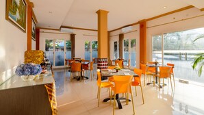 A modern dining area with orange chairs, a glass table, and a view of a body of water through large windows. - The Kanjeng Hotel Kuta (Kuta)