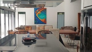 A modern kitchen with a central island, a dining area with a sofa and chairs, and a large window providing natural light. - Casa en Riviera Maya con alberca privada (Puerto Morelos)