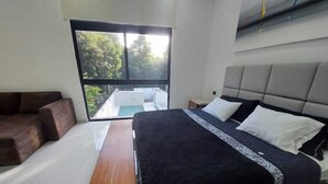 A modern bedroom with a large bed, a bench, a sofa, and a large window offering a view of trees. - Casa en Riviera Maya con alberca privada (Puerto Morelos)