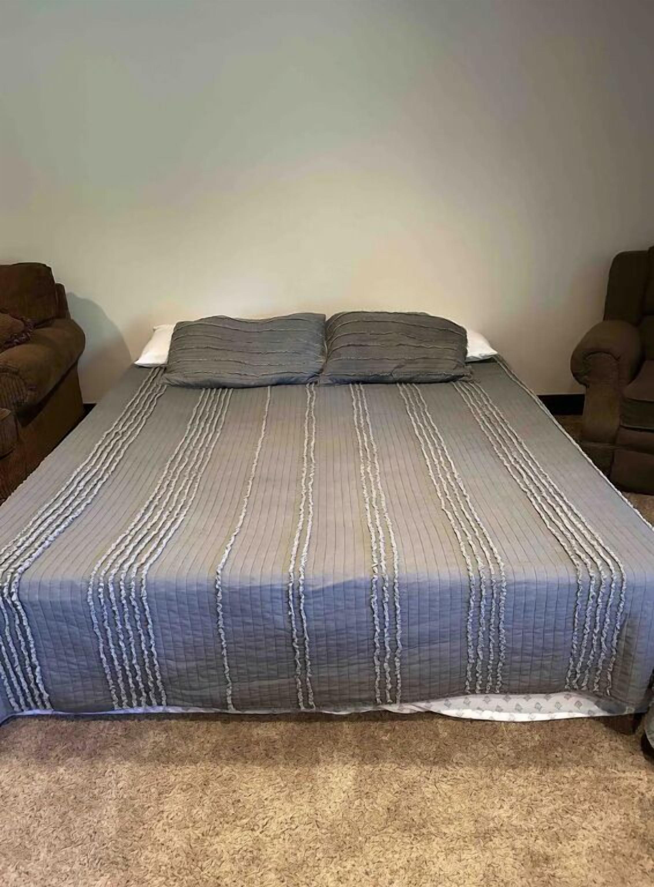 Ein Bett mit gestreifter Bettdecke und zwei Kissen, ein braunes Sofa und ein Sessel in einem Raum mit schlichten Wänden.
