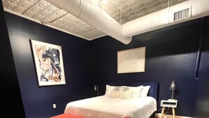 Une chambre à coucher avec un lit, un pouf rouge, une table de chevet et une lampe fixée au mur.