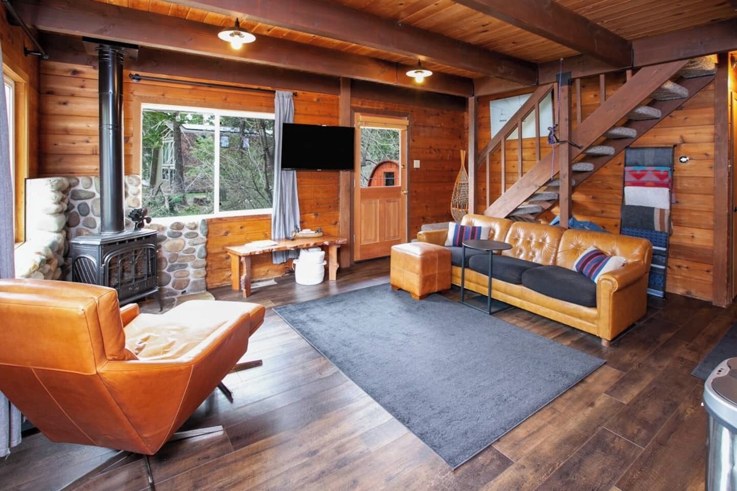A cozy cabin interior with a stone fireplace, leather sofa, and a staircase leading to a loft.