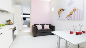 A modern kitchen with white cabinets, a grey sofa, and a white dining table with apples and decorative items. - WHITE HOUSES, Modern and functional studio apartment (bologna)