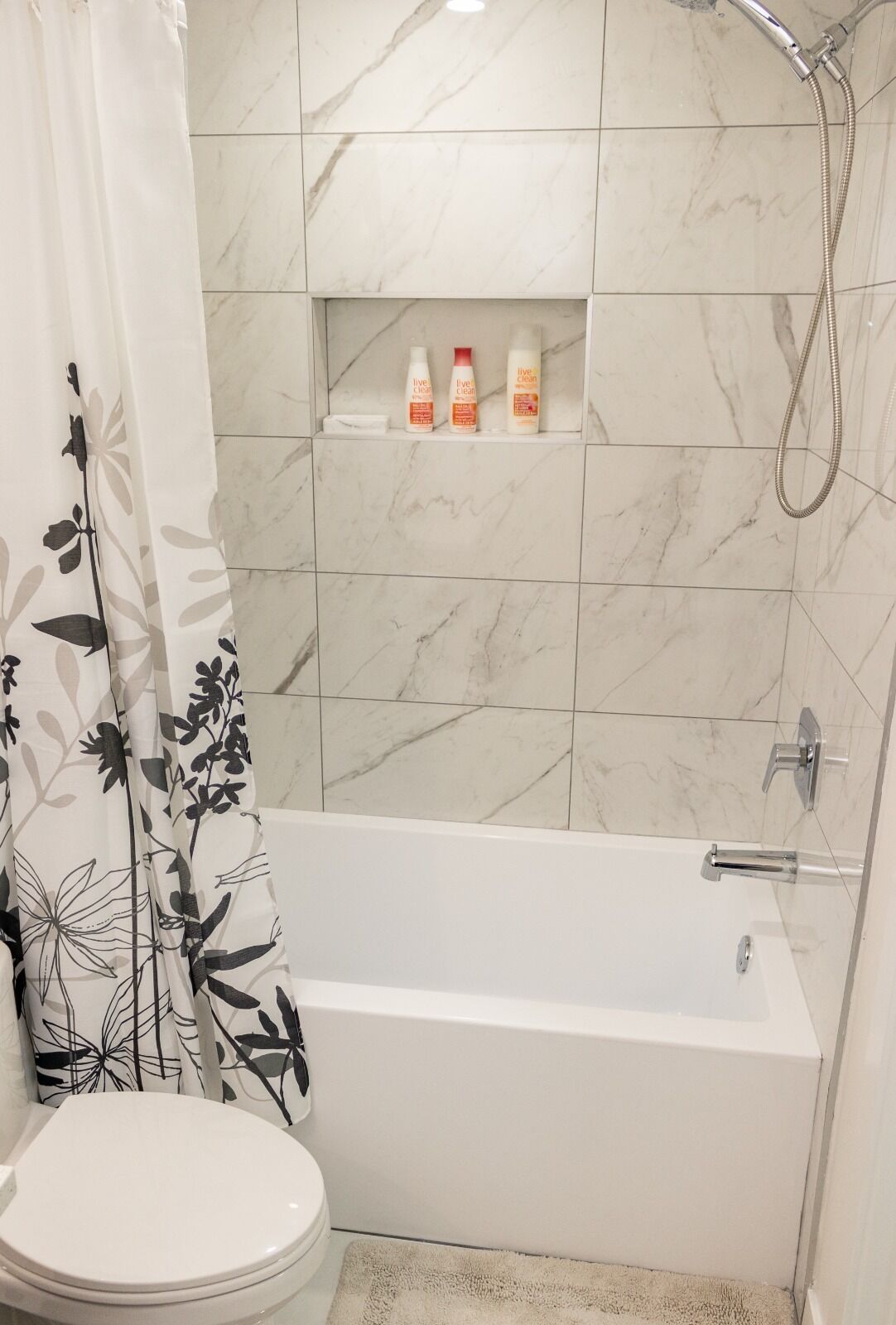 Et badeværelse med et hvidt badekar, toilet og bruseområde. Der er tre flasker på en hylde over badekarret.