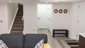A modern living room with a grey sofa, a staircase, a hallway leading to a white door, and a small bench. - Amazing Basement Suite With a Dedicated Workspace (Airdrie)