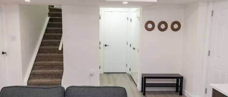 A modern living room with a grey sofa, a staircase, a hallway leading to a white door, and a small bench.