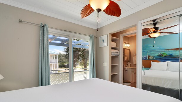 Une chambre dotée d’un grand lit, d’un ventilateur de plafond, d’une vue sur l’extérieur grâce à une porte coulissante, et d’une salle de bain avec une douche et des toilettes visibles.
