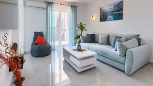 Uma sala de estar moderna com um sofá cinzento, uma mesa de centro branca e uma almofada vermelha. Há uma planta na mesa de centro e um quadro na parede.