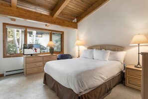Quarto com uma cama grande, duas luminárias de cabeceira, uma TV sobre um aparador de madeira e uma janela com vista para árvores.