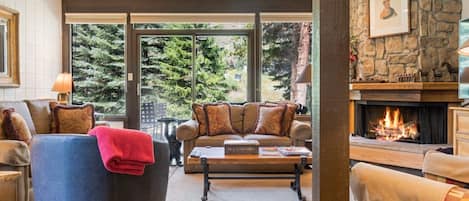 A cozy living room with a fireplace, comfortable seating, and a view of the forest.