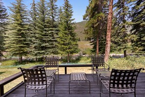A deck with four metal chairs and a table, overlooking a forested area with a river and a hill in the distance.