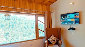 A room with a large window, a wooden ceiling, a wall-mounted TV, and a chair with a patterned cushion. - The Parijat Retreat (Bhikyasain)