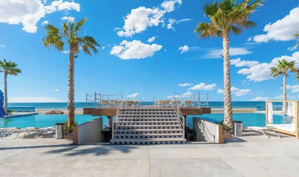 Ein Poolbereich mit Stufen zu einer Terrasse mit Blick auf das Meer, umgeben von Palmen.