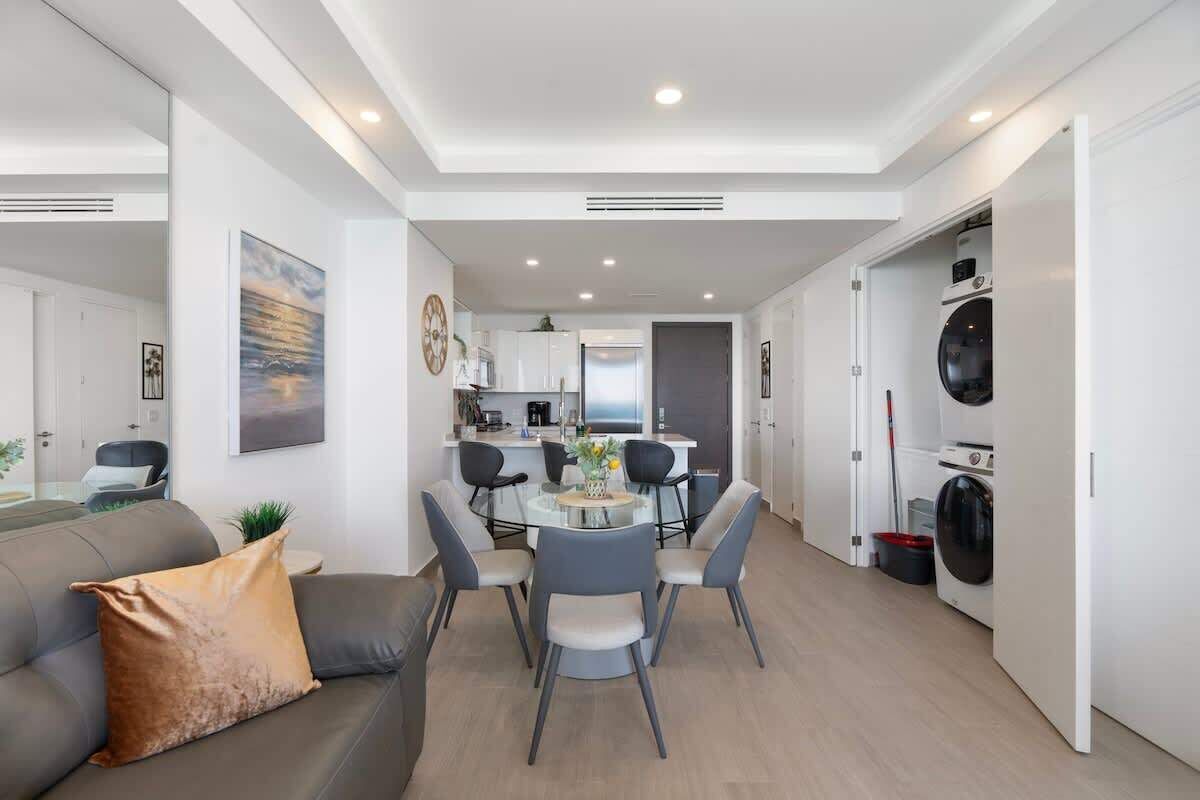 A modern living room with a grey sofa, a dining table with chairs, and a kitchen in the background.