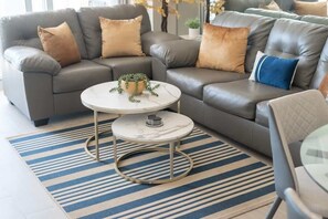 A modern living room with a grey leather sectional sofa, a round coffee table with a marble top, and a blue-striped rug.