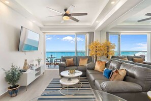 A modern living room with a sofa, coffee table, and TV, overlooking a balcony with a view of the sea.