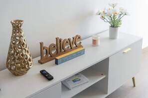 Un tavolino consolle bianco con un vaso decorativo, un telecomando, una candela e un cartello con la scritta 'believe in the moment'.