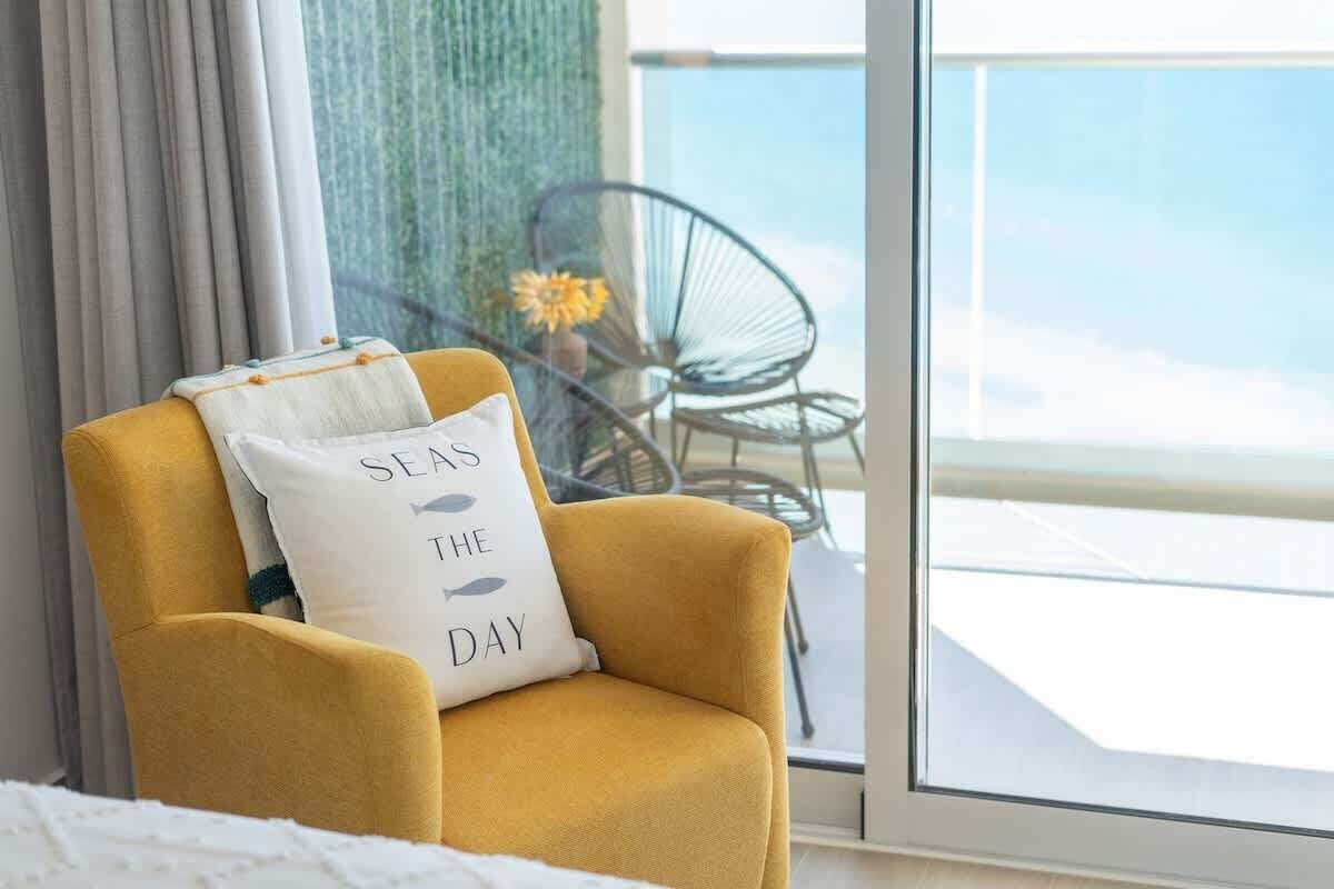 Un fauteuil jaune avec un coussin orné de l’inscription « PROFITE DE LA JOURNÉE » dans une chambre d’hôtel donnant sur la mer grâce à une grande fenêtre.