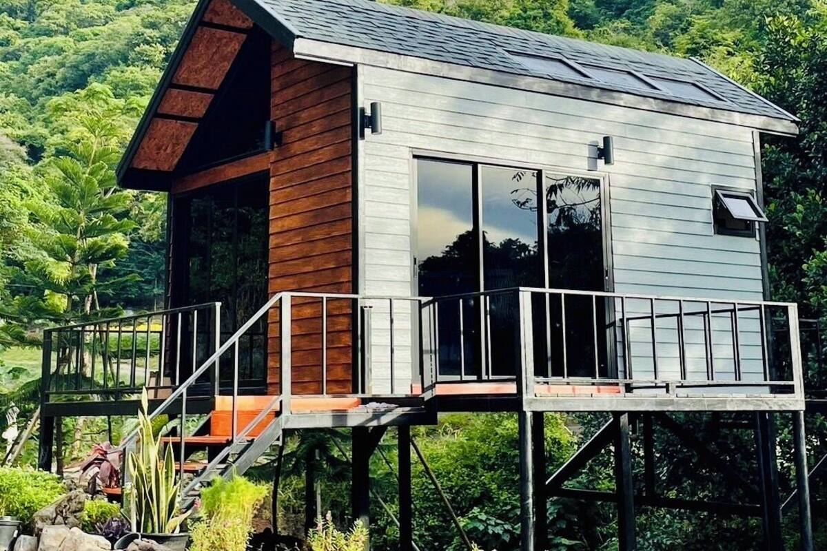 Une maison moderne avec une façade en bois, de grandes fenêtres et un balcon. Elle est construite sur pilotis et offre une vue sur une forêt verdoyante.