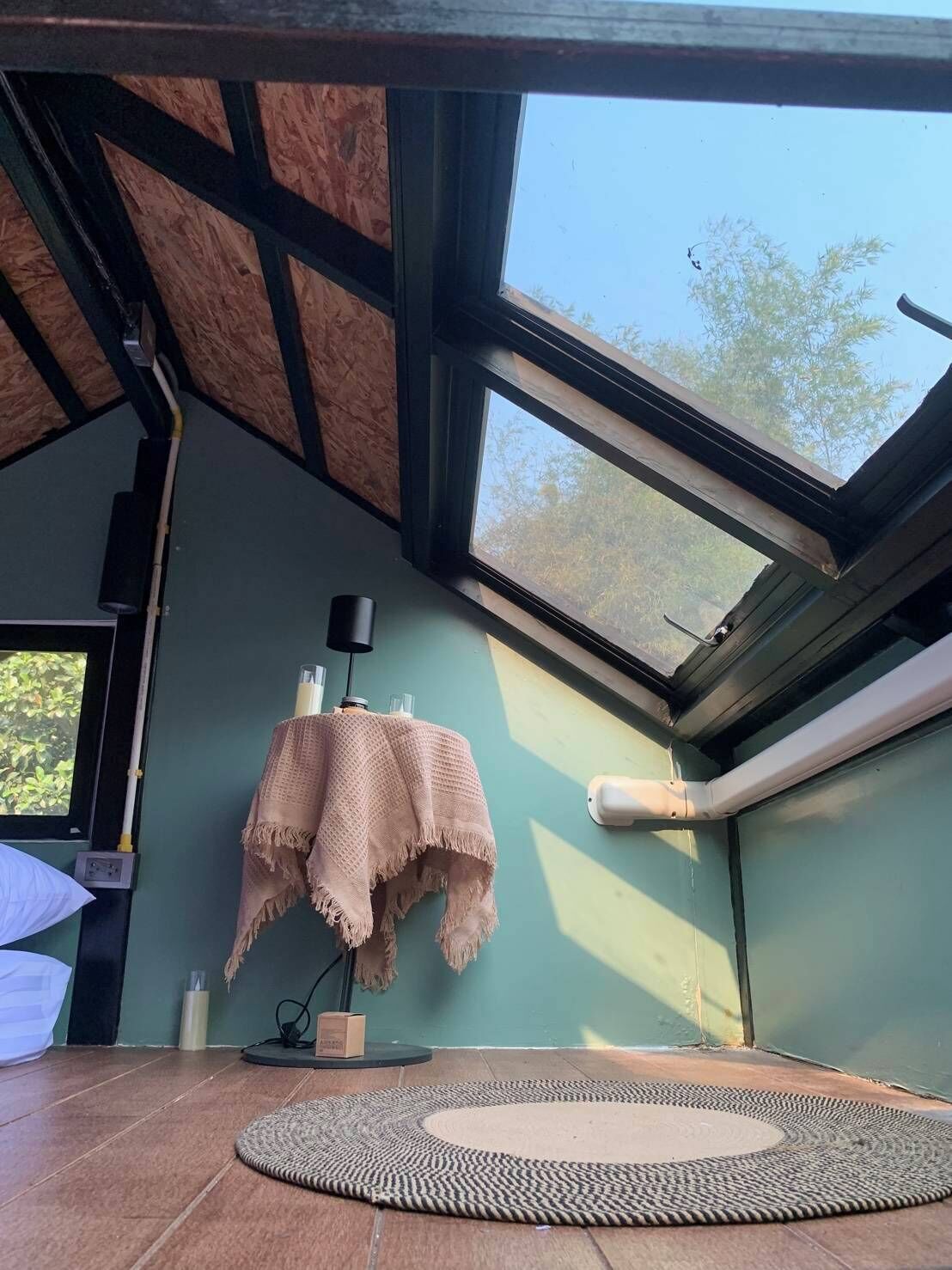 Une pièce avec un plafond en pente, des fenêtres de toit, un tapis rond en fibres naturelles, une petite table recouverte d’une nappe et une chaise.