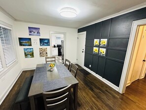 Een eethoek met een lange tafel, stoelen en een bank. Er hangen foto’s aan de muur en is een deur met een zwart paneel.