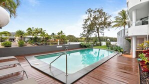 A swimming pool with a wooden deck, surrounded by palm trees and a modern building.