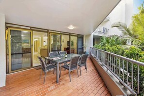 A balcony with a table and chairs, a sliding glass door, and a view of a garden with palm trees.