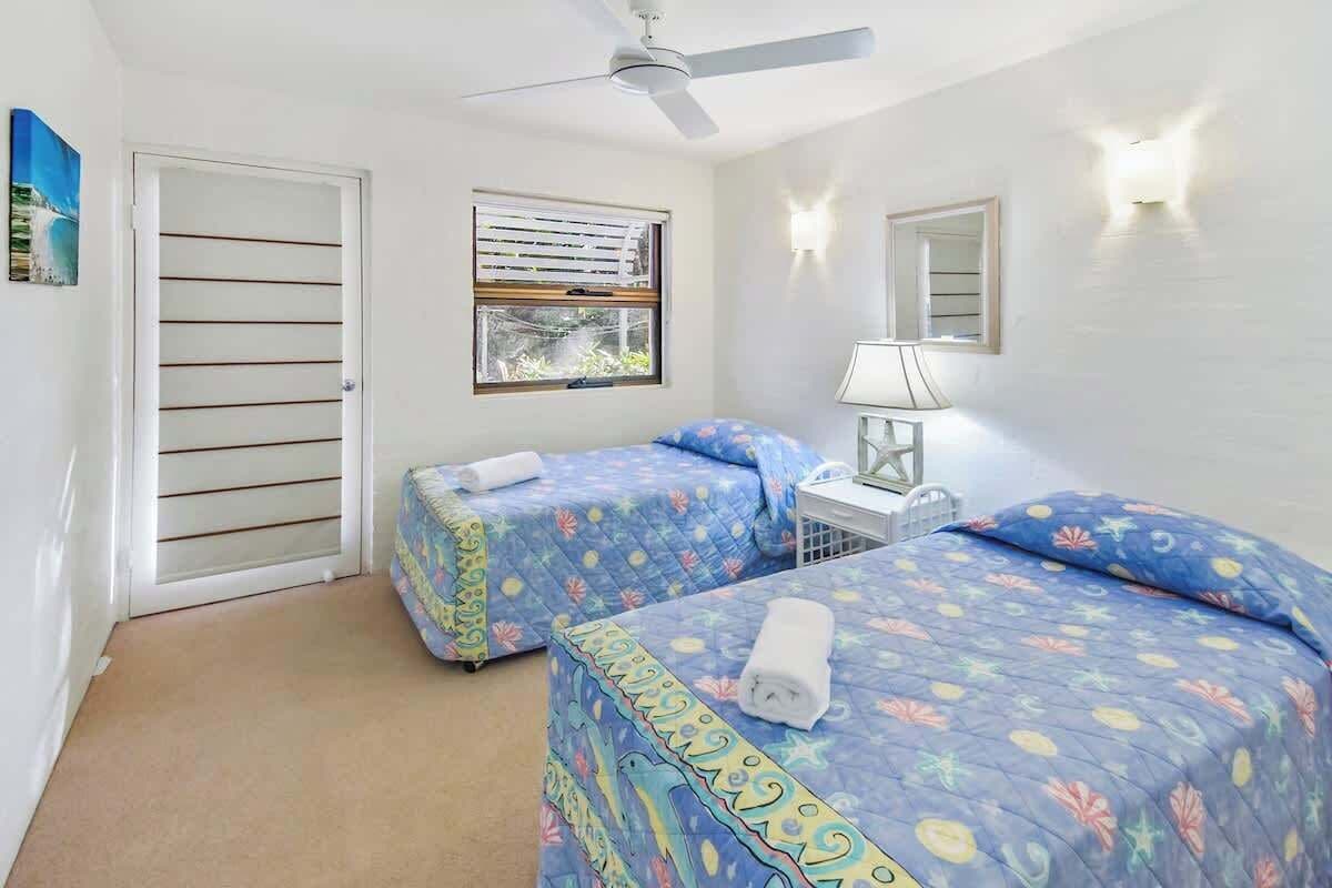 Una habitación con dos camas, un ventilador de techo, una ventana con persianas, una mesita de noche con lámpara y una puerta con persianas horizontales.