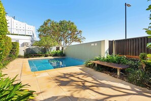 A swimming pool with a wooden bench, surrounded by greenery and a white building.