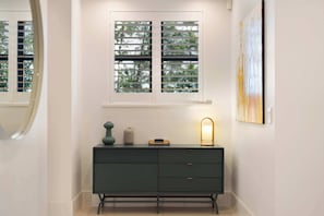 A hallway with a dark green dresser, a window with blinds, and a wall-mounted light fixture.