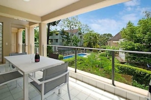 A balcony with a table and chairs, a bottle of wine, and a view of a swimming pool and greenery.