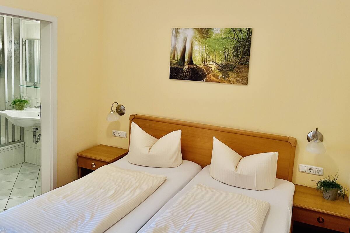 A double bed with white linens, a wooden headboard, bedside tables with lamps, a wall-mounted painting of a forest scene, and a bathroom with a sink and mirror.