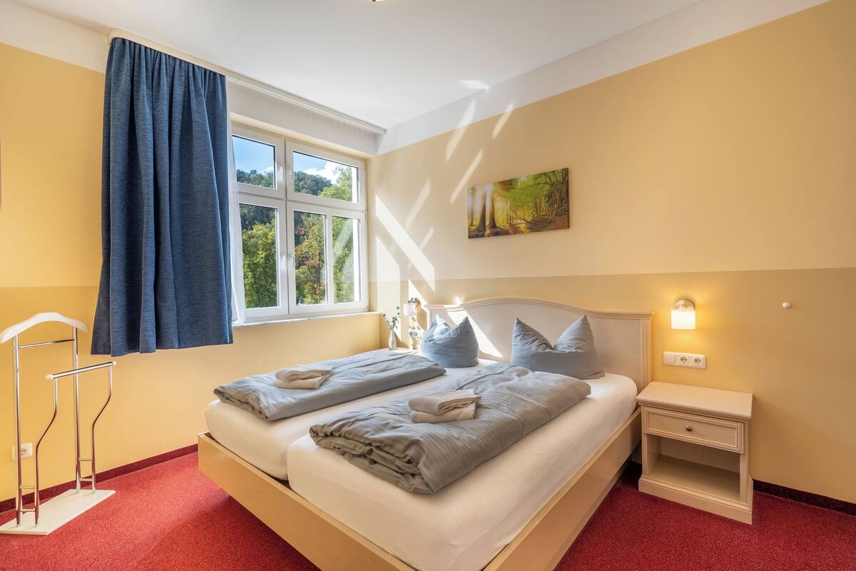 A bedroom with a bed, bedside tables, a window with curtains, a painting on the wall, and a red carpet.
