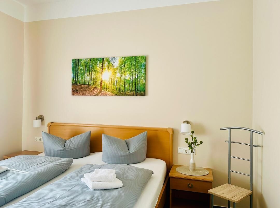 Habitación de hotel con cama, dos almohadas, una mesita de noche, lámpara, un cuadro de un bosque y un estante metálico.