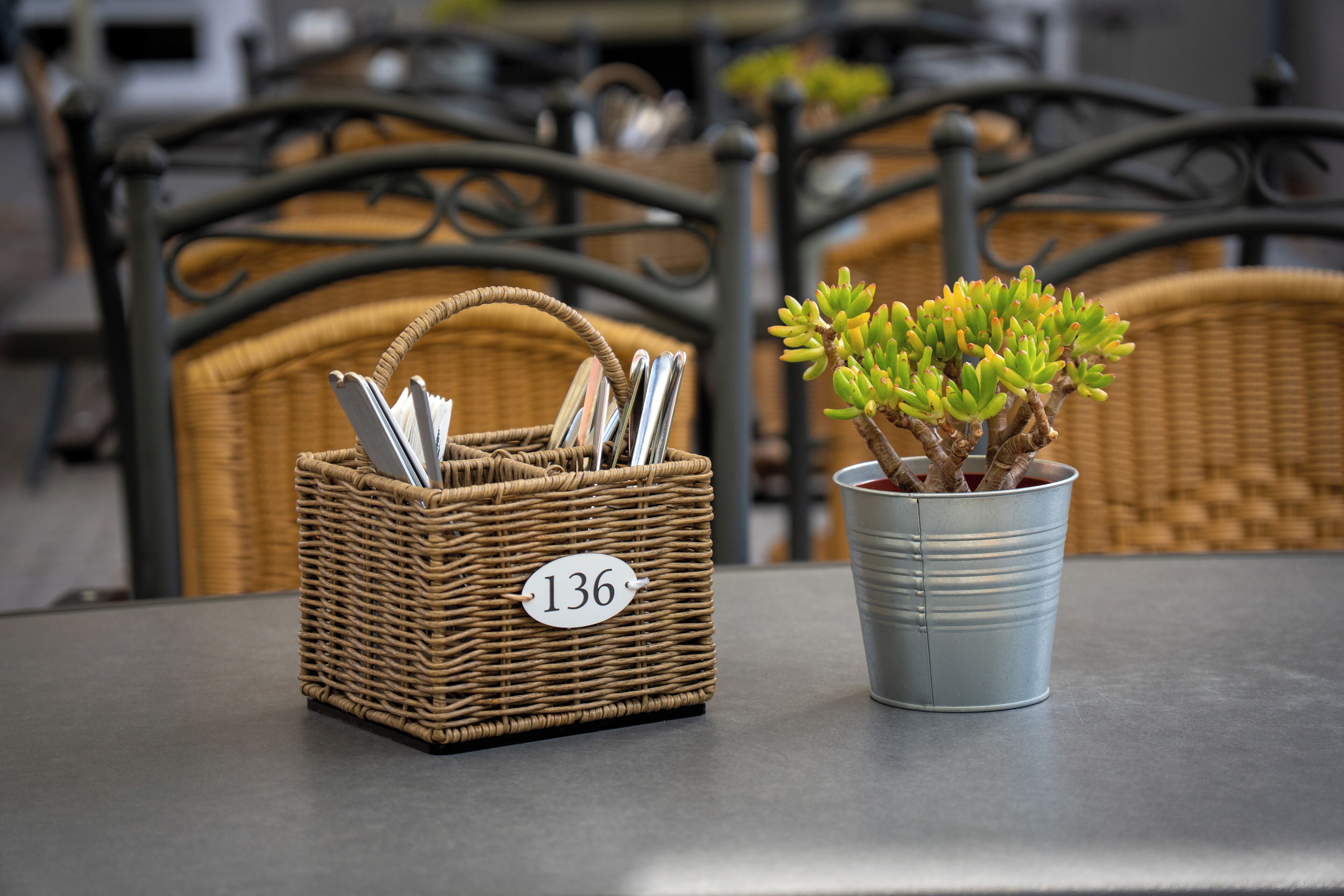 Una cesta de mimbre con la etiqueta '136', cubiertos y una planta en maceta sobre una mesa.