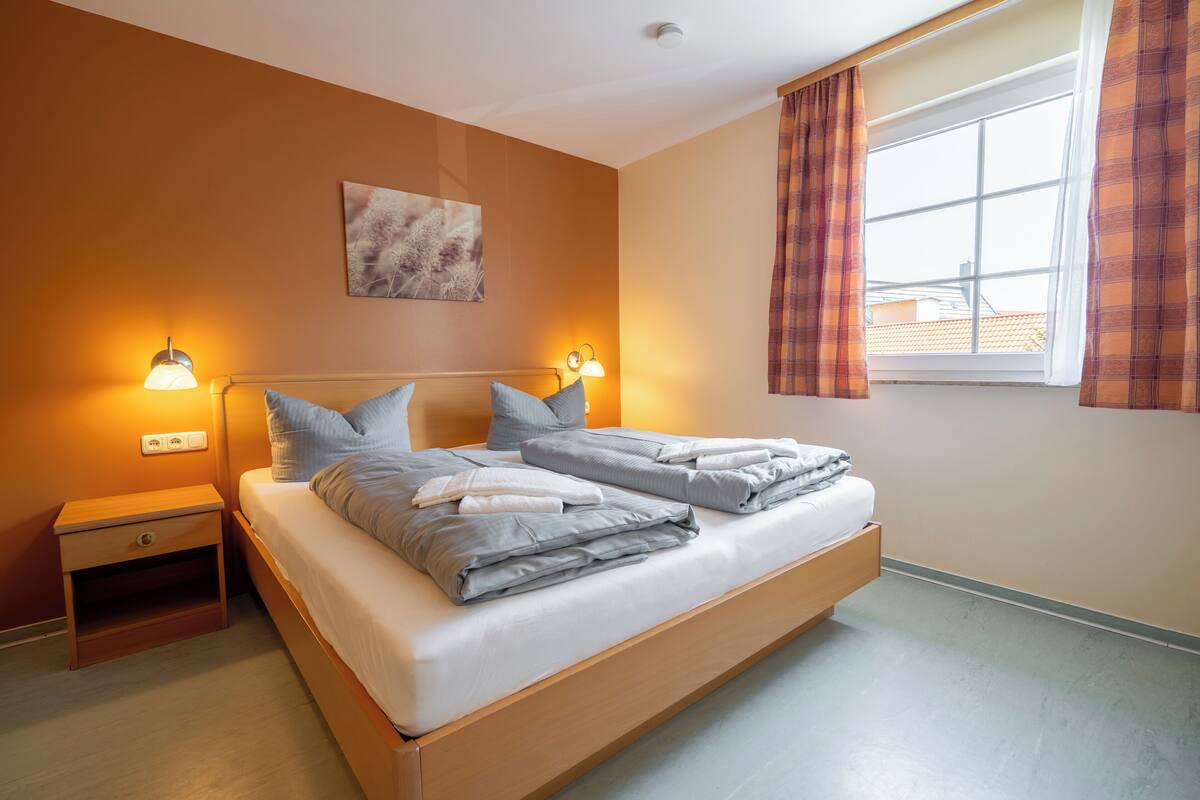 A double bed with grey bedding, two bedside lamps, a small wooden nightstand, a wall-mounted light switch, and a window with plaid curtains.