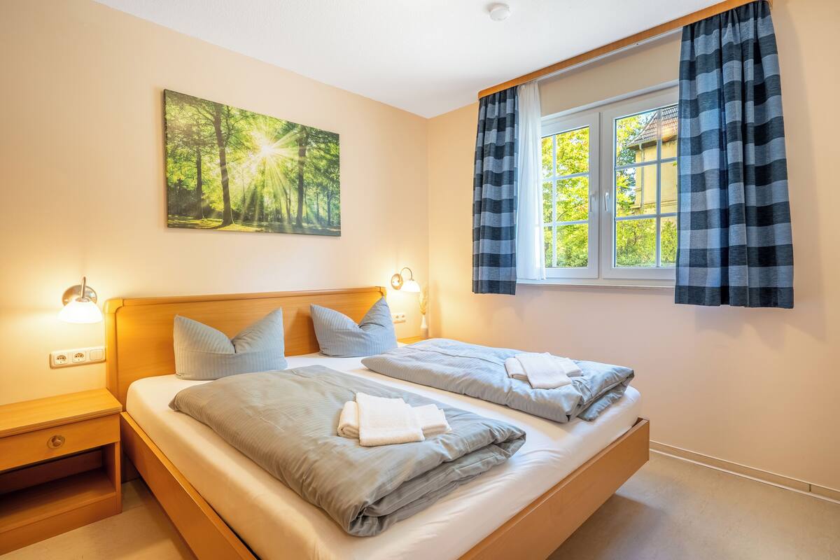 A bedroom with two beds, a window with curtains, a wall-mounted painting, and bedside lamps.