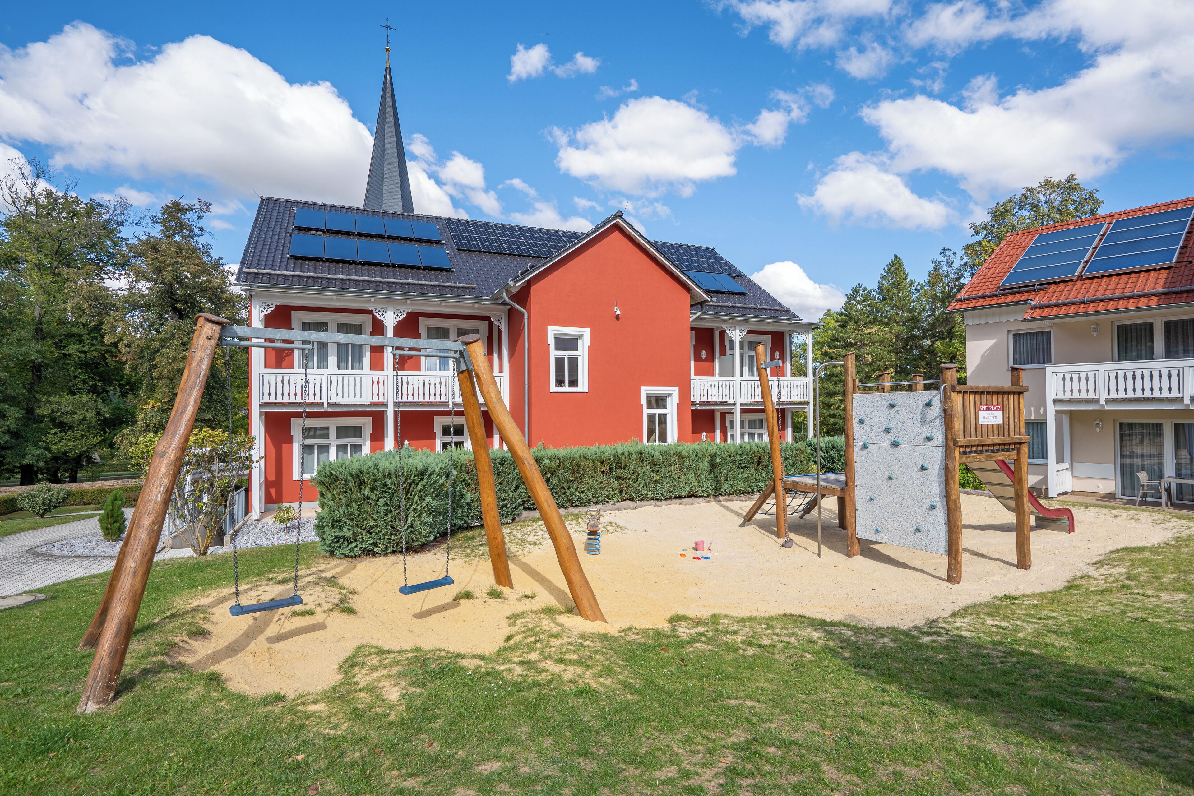 Un parque infantil con columpios y estructuras para escalar, un edificio rojo con campanario de iglesia y paneles solares en el techo.