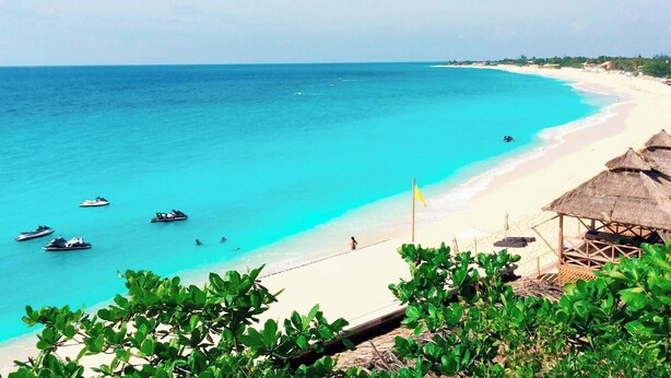 Une plage à l’eau turquoise et limpide, avec un rivage sablonneux et des constructions aux toits de chaume.