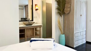 Une chambre d’hôtel avec une coiffeuse en bois, un grand miroir, un lavabo en forme de coupe, un lit avec une serviette blanche et bleue, un vase vert et une armoire en bois.